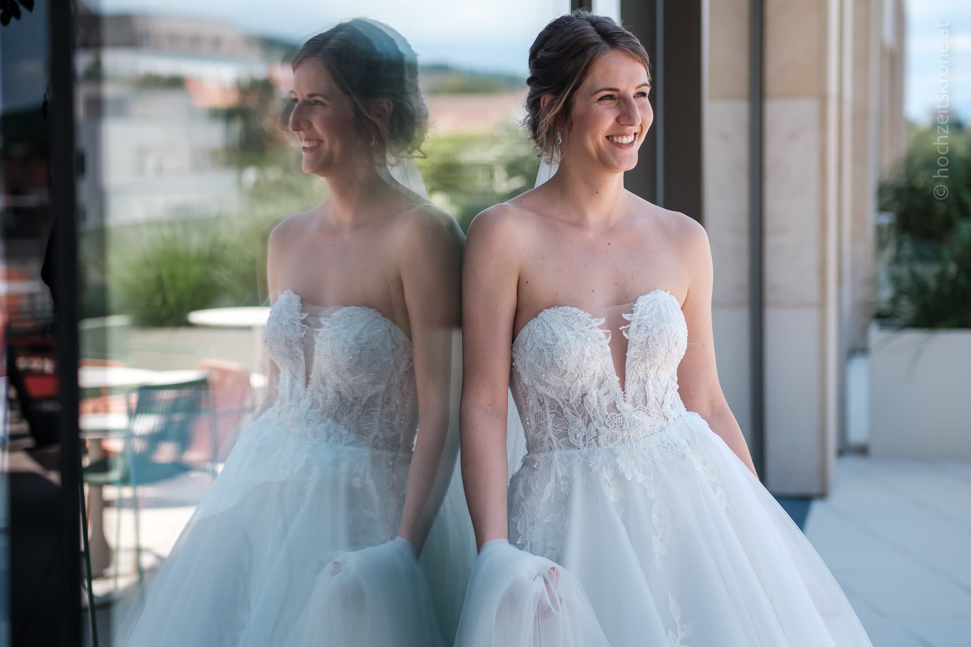 Hochzeitskrone Eine Frau in einem trägerlosen weißen Spitzenbrautkleid steht neben einer reflektierenden Glaswand, lächelt und blickt nach draußen und fängt echte Momente der Freude und Vorfreude ein.