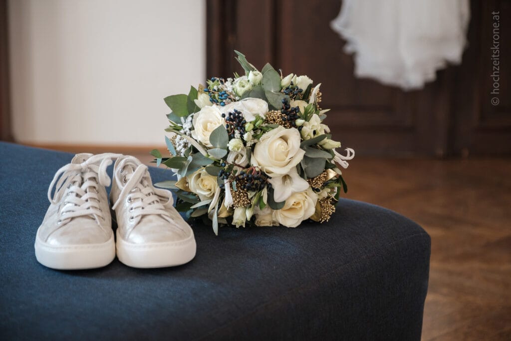 Hochzeitskrone Freizeitschuhe und ein weißer Rosenstrauß ruhen auf einem dunkelblauen Ottomanen, im Hintergrund hängt ein Brautkleid und fängt echte Momente ein, die die Geschichte Ihres besonderen Tages erzählen.