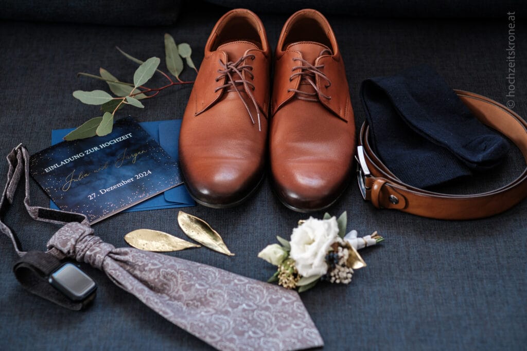 Hochzeitskrone Braune Abendschuhe, schwarze Socken, ein brauner Gürtel, eine gemusterte Krawatte, goldene Blätter, eine Boutonniere, eine Armbanduhr und eine Hochzeitseinladung sind elegant auf einer dunklen Oberfläche arrangiert – so fangen sie echte Momente für authentische Hochzeitsfotos ein.