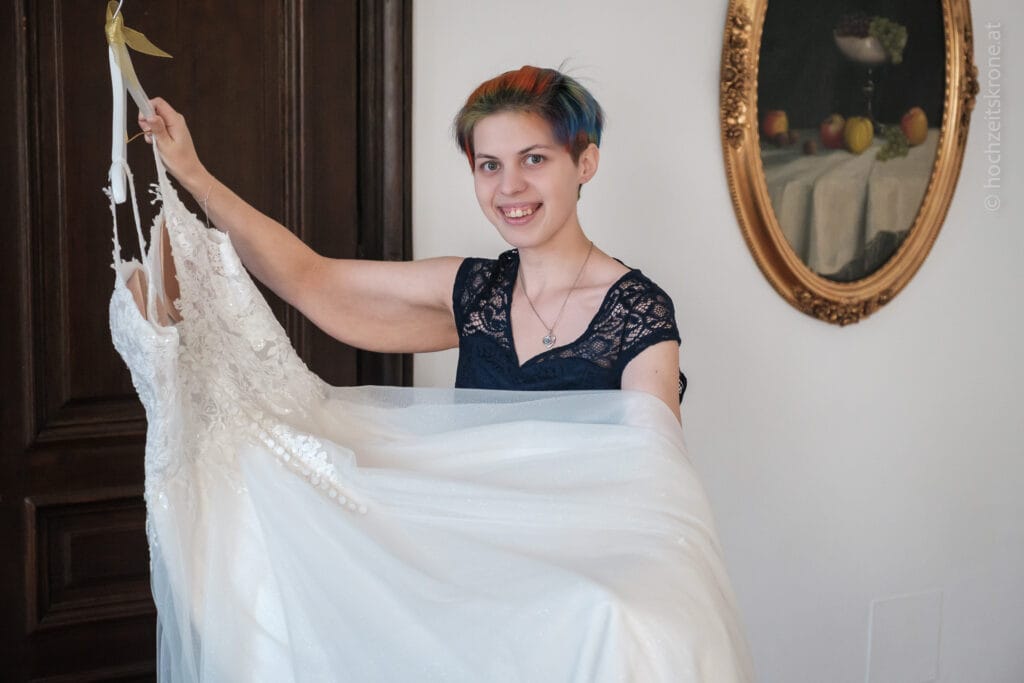 Hochzeitskrone Eine Person strahlt, während sie in einem Raum mit einer Holztür und einem gerahmten Gemälde ein Brautkleid hält und echte Momente festhält; das sind die Art von authentischen Erinnerungen, die Sie von einem Hochzeitsfotografen in Wien und Niederösterreich erwarten würden.