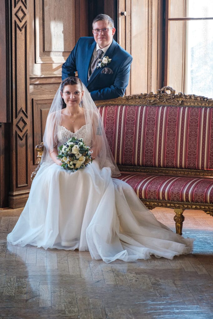 Hochzeitskrone Eine Braut im weißen Kleid sitzt mit einem Blumenstrauß in der Hand auf einem gestreiften Sofa, während ein Bräutigam im blauen Anzug hinter ihr steht. Beide lächeln sich an – ihre echte Freude wurde in einer authentischen Hochzeitsfoto-Session perfekt eingefangen.