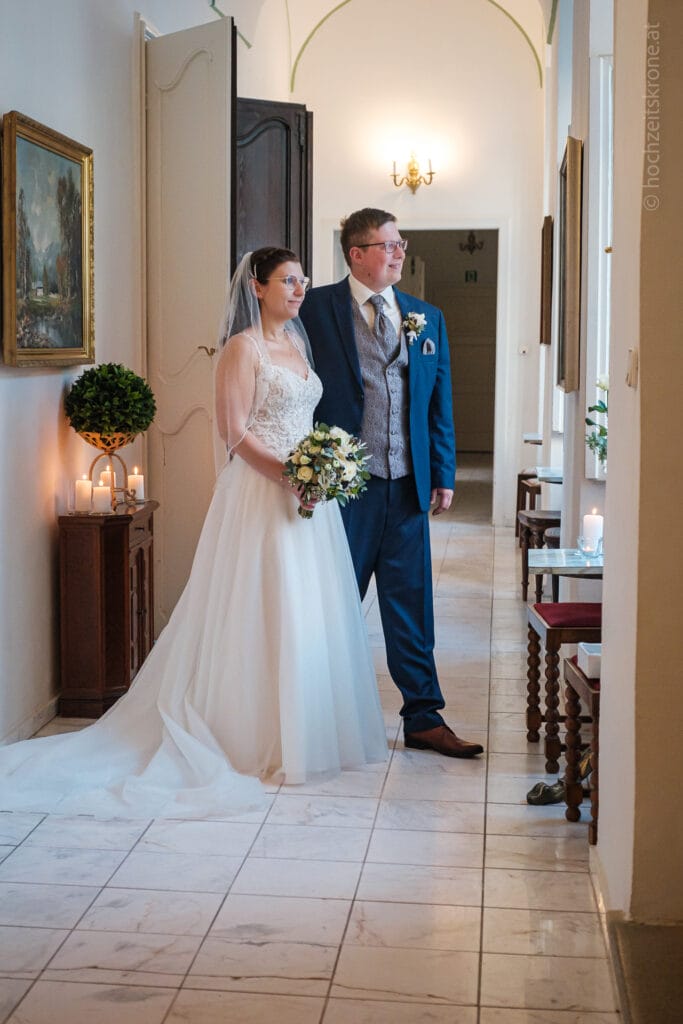 Hochzeitskrone Braut und Bräutigam stehen zusammen in einem Flur, in Hochzeitskleidung gekleidet. Die Braut hält einen Blumenstrauß und auf einem Beistelltisch brennen Kerzen. Diese authentischen Momente, festgehalten von einem Hochzeitsfotografen in Wien und Niederösterreich, spiegeln ihre wahren Gefühle wider.