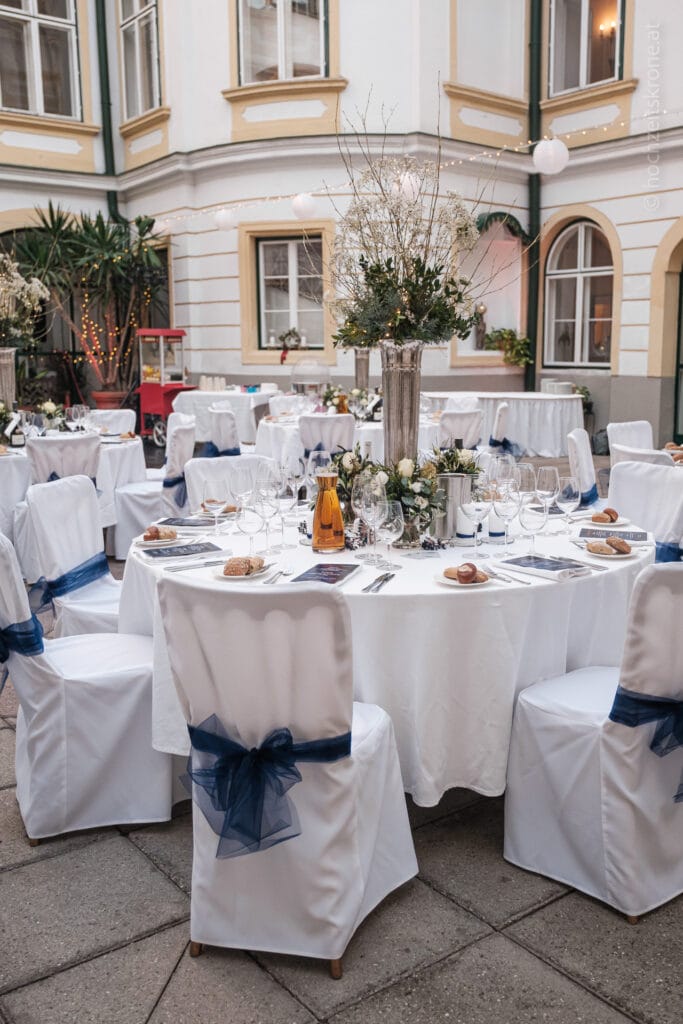 Hochzeitskrone Hochzeitsempfang im Innenhof im Freien mit runden Tischen, die mit weißem Stoff und marineblauen Schleifen bedeckt sind und mit elegantem Geschirr, floralen Tafelaufsätzen und Weinflaschen gedeckt sind. Halten Sie echte Momente fest und bewahren Sie sie für immer mit authentischen Hochzeitsfotos von einem erstklassigen Hochzeitsfotografen in Wien und Niederösterreich.