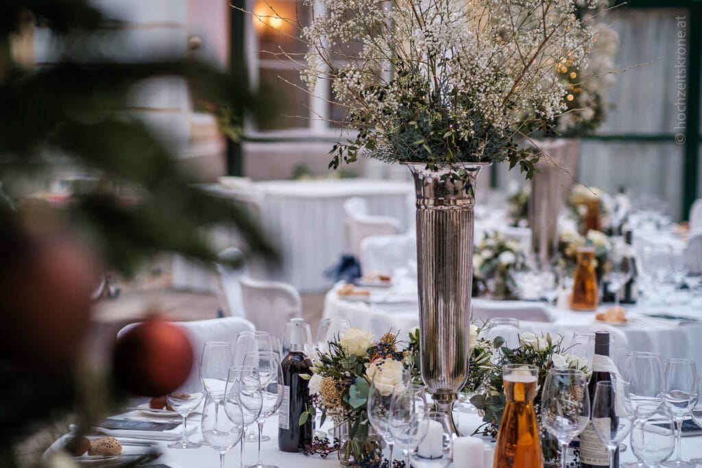 Hochzeitskrone Elegante Tischdekoration mit weißen Tischdecken, hohen silbernen Vasen mit Blumen und verschiedenen Weinflaschen. Gläser und Besteck sind ordentlich angeordnet, perfekt, um echte Momente festzuhalten. Ideal für einen Hochzeitsfotografen in Wien und Niederösterreich, der natürliche Hochzeitsfotos sucht.