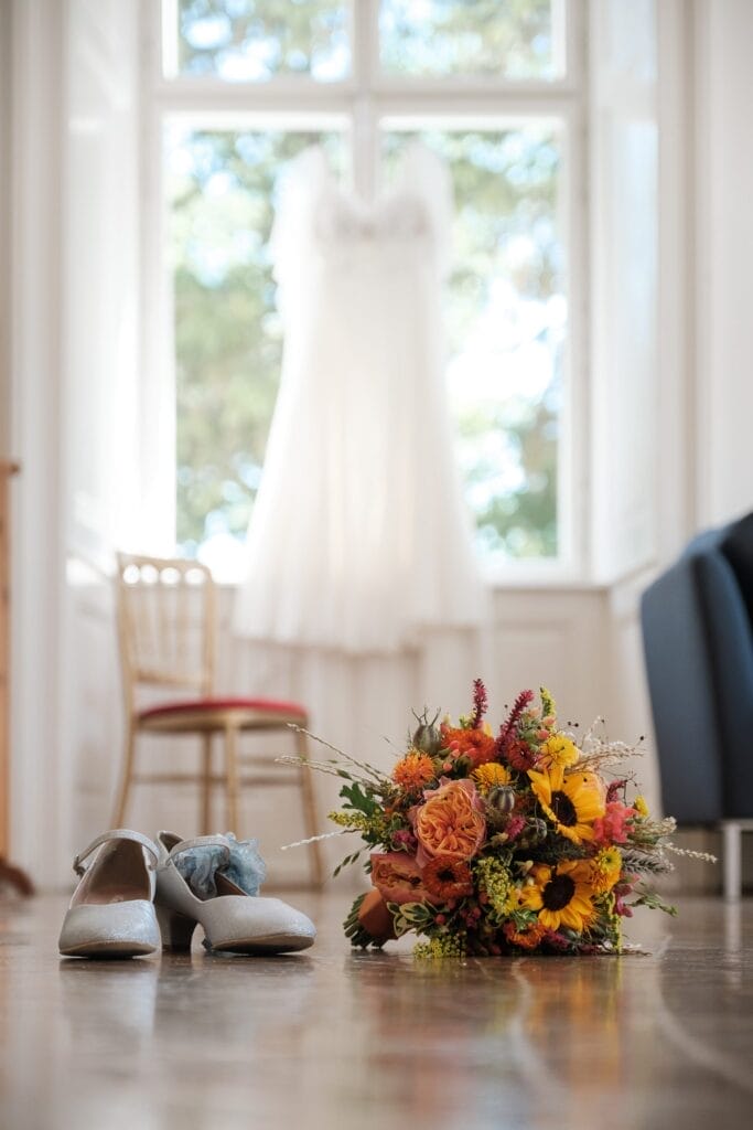 Hochzeitskrone Ein Brautkleid hängt an einem Fenster und fängt echte Momente der Vorfreude ein; ein Blumenstrauß und weiße Schuhe sind elegant auf dem Holzboden platziert und schaffen eine Atmosphäre, die nach authentischen Hochzeitsfotos verlangt.