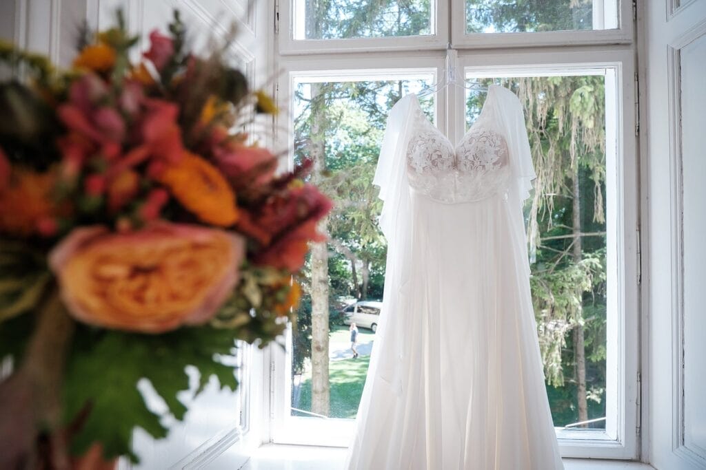 Hochzeitskrone Ein weißes Brautkleid hängt vor einem großen Fenster mit Blick auf üppige Bäume, perfekt eingefangen von einem Hochzeitsfotografen in Wien und Niederösterreich. Im Vordergrund trägt ein Strauß leuchtender Blumen zum Charme dieser natürlichen Hochzeitsfotos bei.