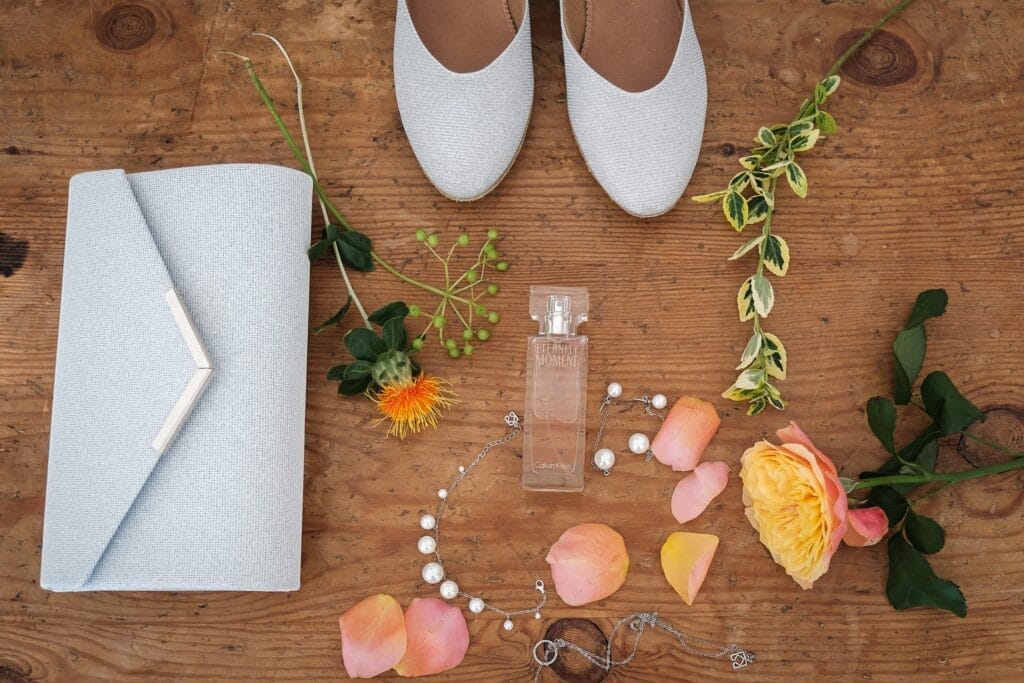 Hochzeitskrone Flache Lage aus weißen Schuhen, passender Clutch, Parfümflasche, Blumen, Perlenkette und silbernem Armband auf einer Holzoberfläche – perfekt, um echte Momente in authentischen Hochzeitsfotos von einem Hochzeitsfotografen in Wien und Niederösterreich einzufangen.