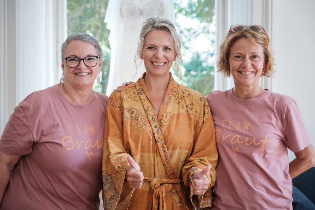 Hochzeitskrone Drei strahlende Frauen; die Frau in der Mitte trägt eine orangefarbene Robe, flankiert von zwei Frauen in „Team Braut“-T-Shirts. Sie werden von einem Fenster mit üppigem Grün davor eingerahmt und fangen die Essenz authentischer Hochzeitsfotos ein.