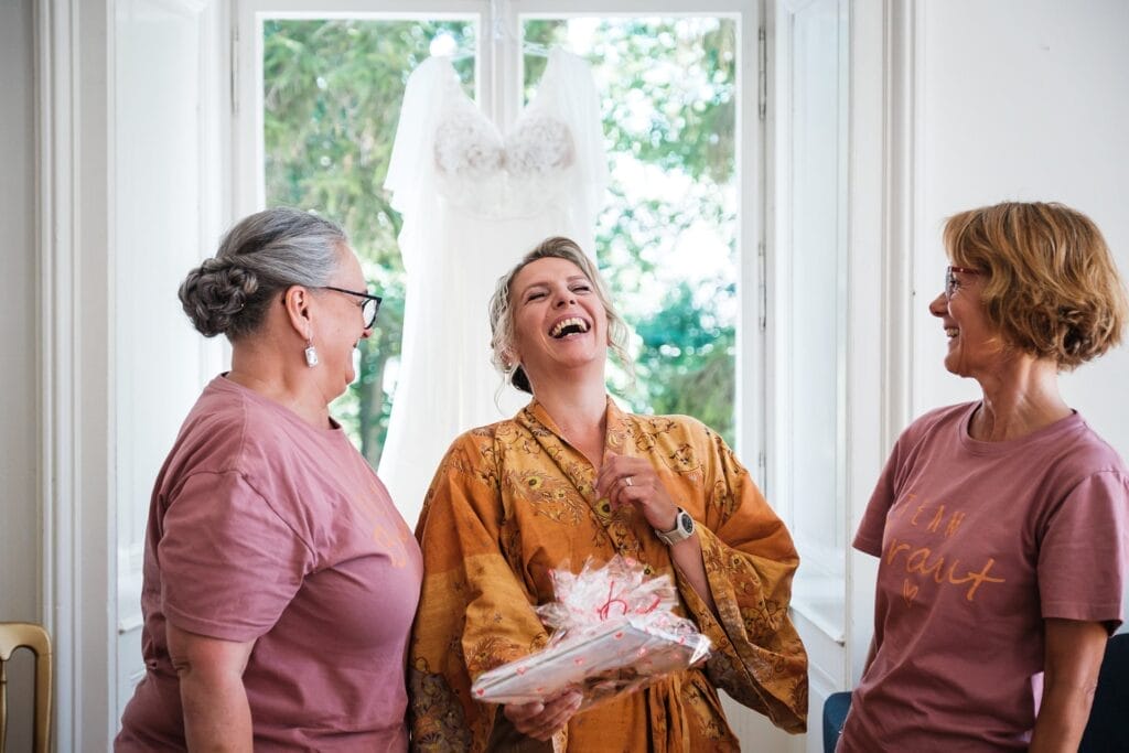 Hochzeitskrone Drei Frauen lachen vor einem Fenster, im Hintergrund hängt ein Brautkleid – authentische Hochzeitsfotos, die echte Momente einfangen.