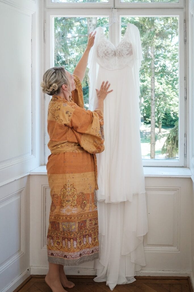 Hochzeitskrone Eine Frau in einem gemusterten Morgenmantel steht an einem Fenster und streckt die Hand aus, um ein langes weißes Kleid zu richten, das neben ihr hängt. So fängt sie echte Momente ein, die sich für natürliche Hochzeitsfotos eignen.