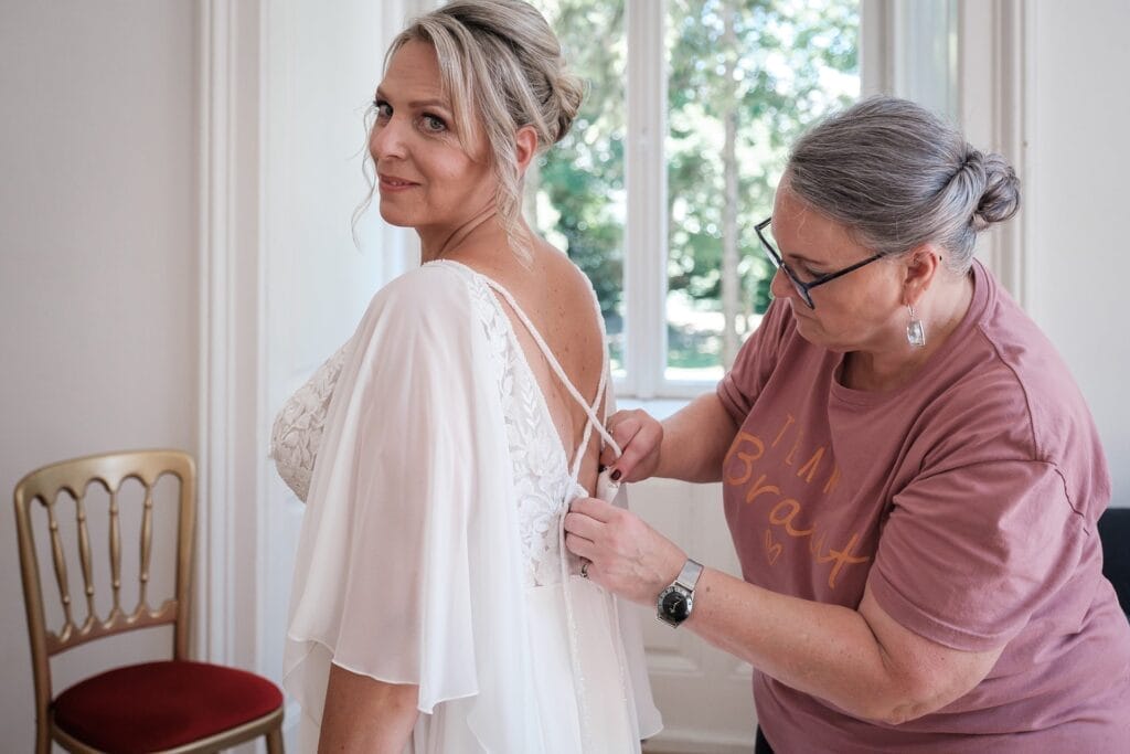 Hochzeitskrone Eine Frau in einem weißen Kleid lässt sich von einer anderen Frau in einem rosa Hemd den Rücken richten und fängt echte Momente ein. Sie befinden sich in einem Raum mit großen Fenstern, die einen ruhigen Blick auf die Bäume draußen bieten, was an natürliche Hochzeitsfotos erinnert.