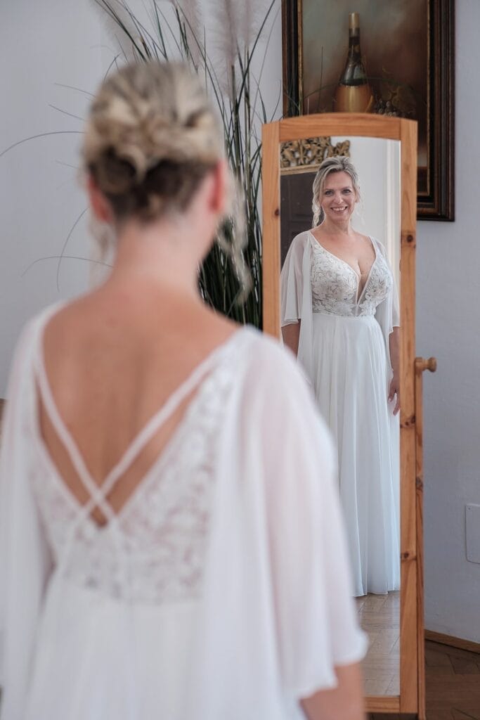 Hochzeitskrone Eine Frau in einem weißen Kleid mit Spitzendetails auf dem Rücken steht vor einem Spiegel, der ihr lächelndes Bild reflektiert. Echte Momente werden festgehalten, indem eine Pflanze und ein gerahmtes Bild ruhig im Hintergrund stehen.