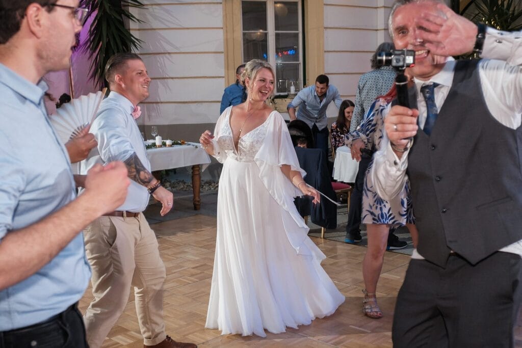 Hochzeitskrone Bei einem Hochzeitsempfang im Innenbereich strahlt eine Braut in einem weißen Kleid in der Mitte, umgeben von tanzenden Gästen. Ein Mann in einer Weste, wahrscheinlich der Hochzeitsfotograf in Wien und Niederösterreich, fängt authentische Hochzeitsfotos ein und hält diese echten Momente für immer fest.