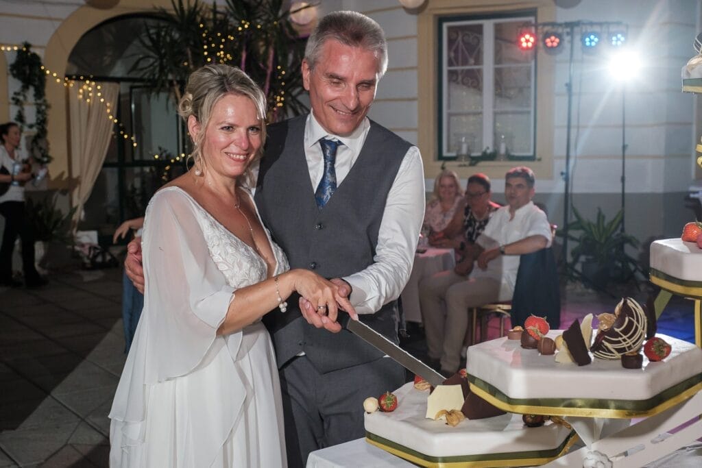 Hochzeitskrone Ein Paar teilt echte Momente, als es bei seiner Feier in geschlossenen Räumen eine mehrstöckige Hochzeitstorte anschneidet, die mit Obst und Schokolade geschmückt ist. Perfekt festgehalten von einem Hochzeitsfotografen in Wien und Niederösterreich. Im Hintergrund sitzende Gäste erfreuen sich an diesen natürlichen Hochzeitsfotos.