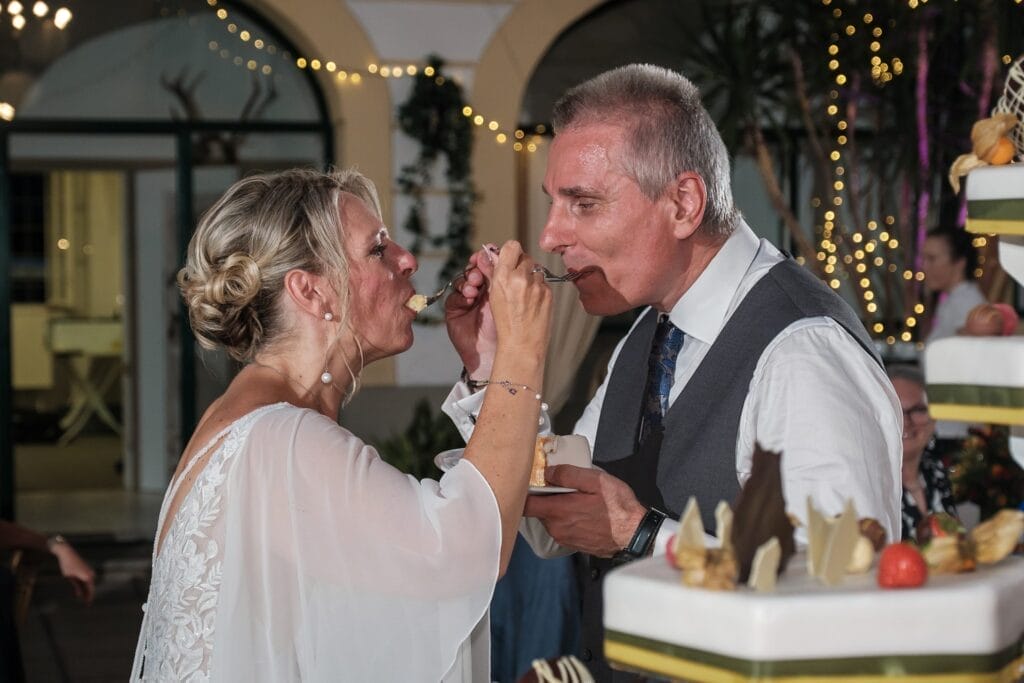 Hochzeitskrone Bei einem eleganten Treffen teilt sich ein Paar ein Stück Kuchen und füttert sich gegenseitig zärtlich mit Gabeln. Eine dreistöckige Torte ziert die Szene und fängt echte Momente ein, die an authentische Hochzeitsfotos eines talentierten Hochzeitsfotografen aus Wien und Niederösterreich erinnern.