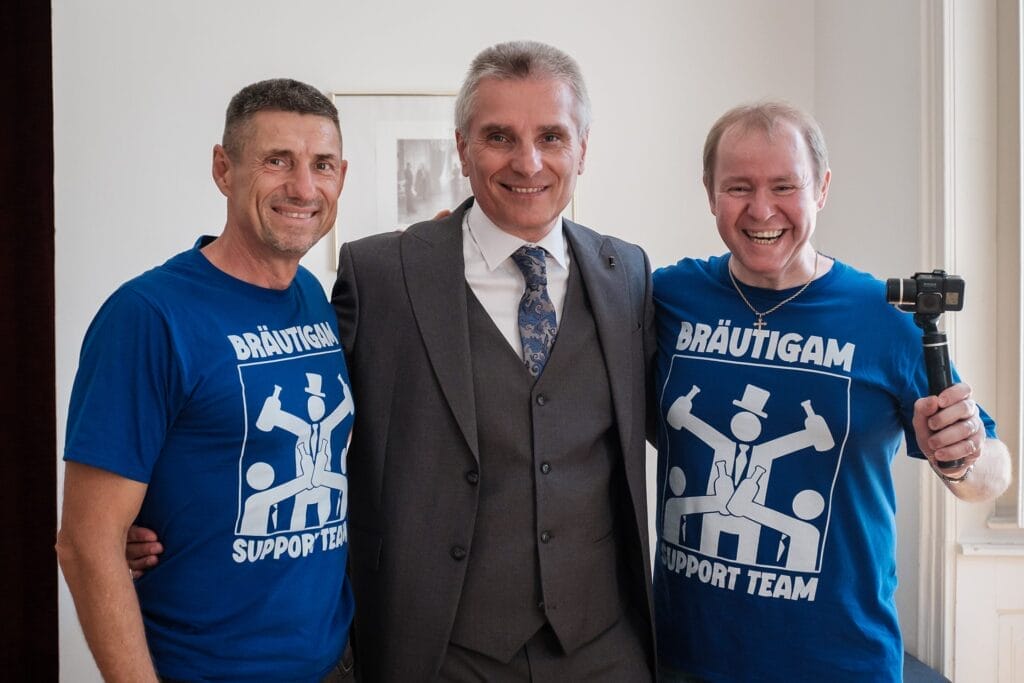 Hochzeitskrone Drinnen lächeln drei Männer; Der Mann in der Mitte trägt einen Anzug, während seine Begleiter blaue „Bräutigam Support Team“-T-Shirts tragen. Diese echten Momente fangen die Freude und Kameradschaft ein, perfekt für jeden Hochzeitsfotografen in Wien und Niederösterreich, um authentische Hochzeitsfotos zu dokumentieren.