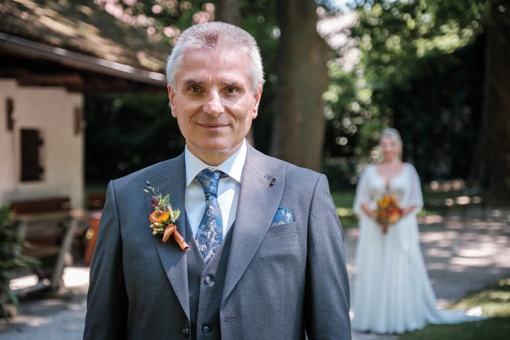 Hochzeitskrone Ein Mann im Anzug mit geblümter Ansteckblume steht im Vordergrund und fängt echte Momente ein, während eine Frau im Hochzeitskleid einen Blumenstrauß in den verschwommenen Hintergrund im Freien hält – perfekt für natürliche Hochzeitsfotos von jedem Hochzeitsfotografen in Wien und Niederösterreich.