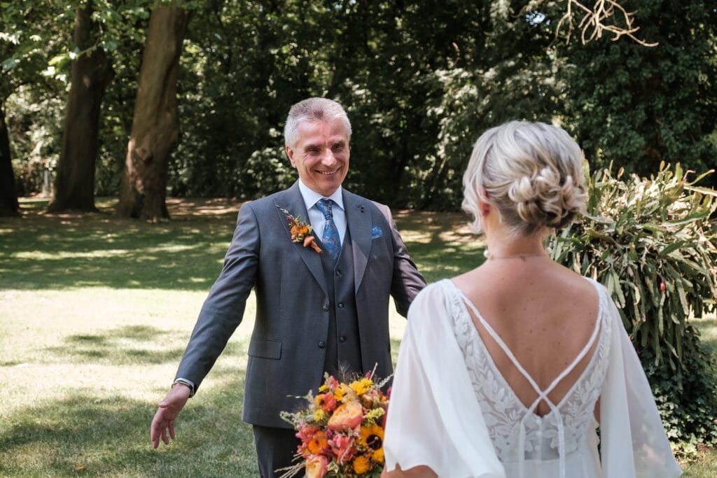 Hochzeitskrone Ein Mann im Anzug lächelt mit offenen Armen, während ihm unter den Bäumen eine Frau in einem weißen Kleid mit einem Blumenstrauß entgegentritt. Diese natürlichen Hochzeitsfotos wurden von einem Hochzeitsfotografen in Wien und Niederösterreich aufgenommen und spiegeln auf wunderbare Weise ihre echten Momente wider.