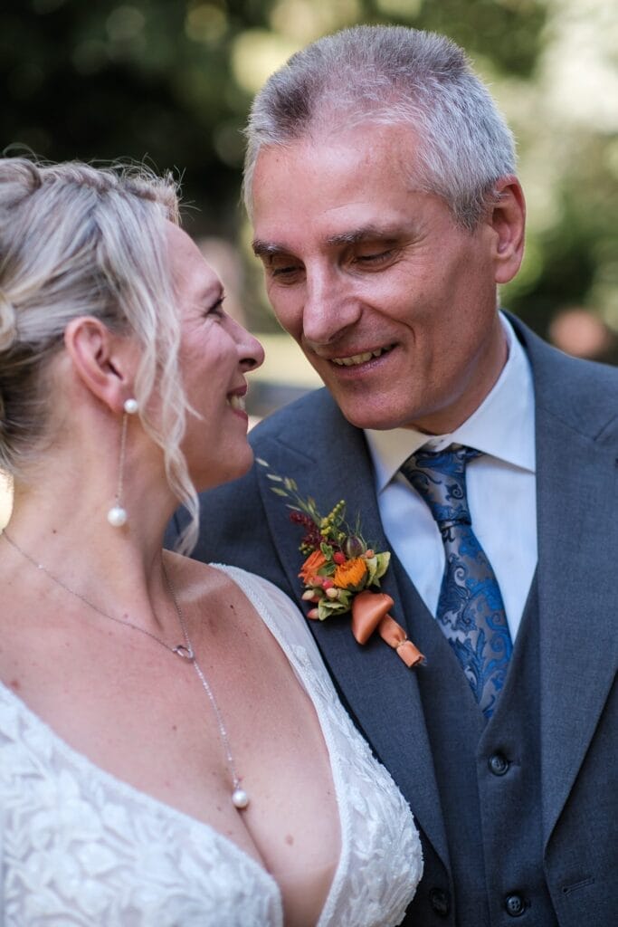 Hochzeitskrone Ein Paar lächelt sich im Freien an; die Frau im weißen Kleid und der Mann im grauen Anzug mit Blumenboutonniere. Diese natürlichen Hochzeitsfotos fangen ihre echte Freude perfekt ein, wie es ein Hochzeitsfotograf in Wien und Niederösterreich meisterhaft geschafft hat.