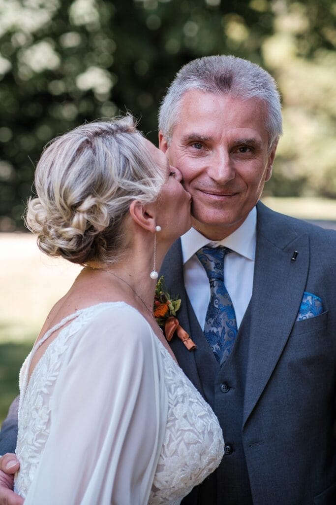 Hochzeitskrone Eine Frau im weißen Kleid küsst einen Mann im grauen Anzug im Freien auf die Wange und fängt damit echte Momente ihres Tages ein.