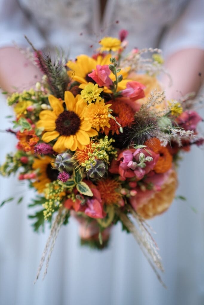 Hochzeitskrone Eine Person hält einen farbenfrohen Blumenstrauß mit Sonnenblumen, rosa Rosen und verschiedenen Wildblumen mit Grünpflanzen und fängt die Essenz echter Momente ein, perfekt für authentische Hochzeitsfotos.