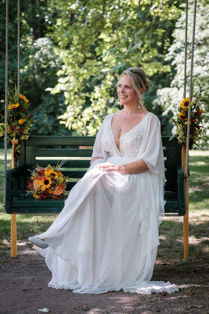 Hochzeitskrone Eine Person in einem weißen Kleid sitzt auf einer grünen, mit Blumen geschmückten Schaukel, hält einen Blumenstrauß und fängt echte Momente ein. Der Hintergrund besteht aus üppig grünen Bäumen, perfekt für natürliche Hochzeitsfotos von einem Hochzeitsfotografen in Wien und Niederösterreich.