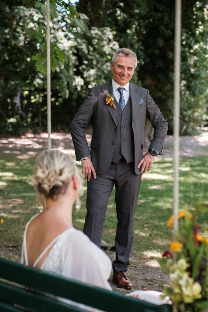 Hochzeitskrone Ein Mann im grauen Dreiteiler lächelt unter einem Baum, während ihm eine sitzende Frau im weißen Kleid gegenübersteht, umgeben von Blumen – eine authentische Hochzeitsfoto-Kulisse, die echte Momente der Freude einfängt.