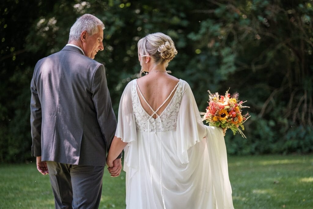 Hochzeitskrone Ein Paar geht Hand in Hand, die Frau hält einen Strauß bunter Blumen, festgehalten in authentischen Hochzeitsfotos inmitten üppigen Grüns.