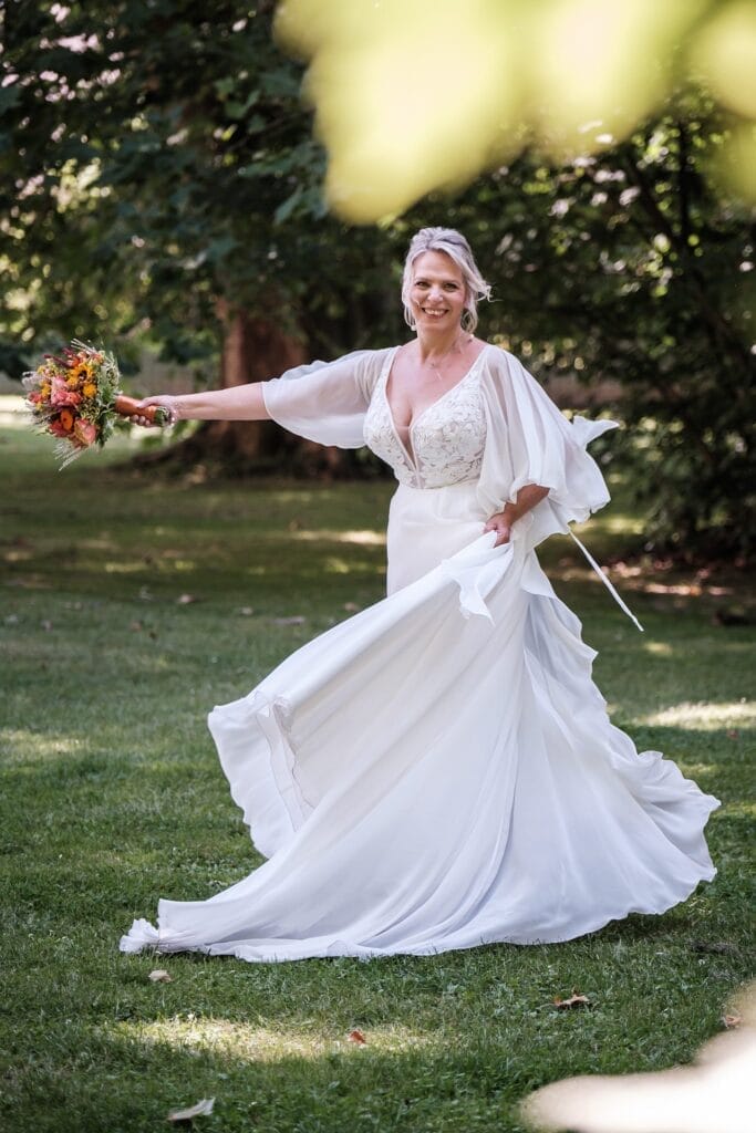 Hochzeitskrone Eine Braut in einem fließenden weißen Kleid hält einen Strauß orangefarbener Blumen und verkörpert echte Momente, während sie auf Gras mit Bäumen im Hintergrund steht und natürliche Hochzeitsfotos macht.