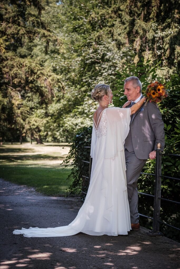 Hochzeitskrone Ein Paar steht auf einem Weg im Grünen und fängt echte Momente ein. Die Frau im weißen Kleid hält einen Strauß Sonnenblumen in der Hand, blickt auf den Mann im Anzug, der an einem Geländer lehnt und natürliche Hochzeitsfotos verkörpert.