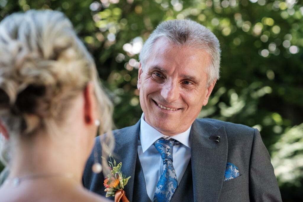 Hochzeitskrone Ein Mann im Anzug mit geblümter Boutonniere lächelt eine Frau mit blonden Haaren im Freien an und fängt echte Momente ein, die ein Hochzeitsfotograf in Wien und Niederösterreich schätzen würde.