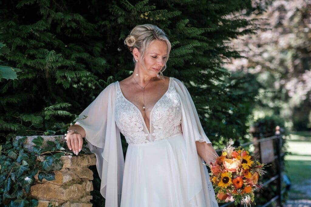 Hochzeitskrone Eine Braut im weißen Kleid steht im Freien, hält einen Strauß gelber und orangefarbener Blumen, ist von Grün umgeben und macht authentische Hochzeitsfotos.