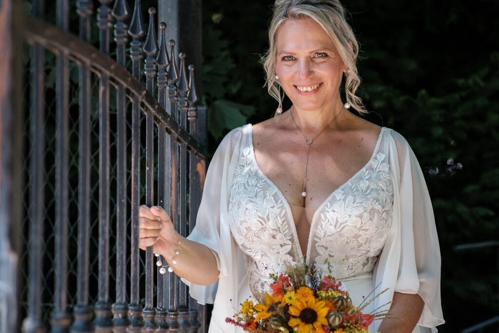 Hochzeitskrone Eine Frau in einem weißen Kleid steht mit einem Blumenstrauß in der Hand lächelnd neben einem schmiedeeisernen Tor; ein perfekter Moment, festgehalten von einem Hochzeitsfotografen in Wien und Niederösterreich.