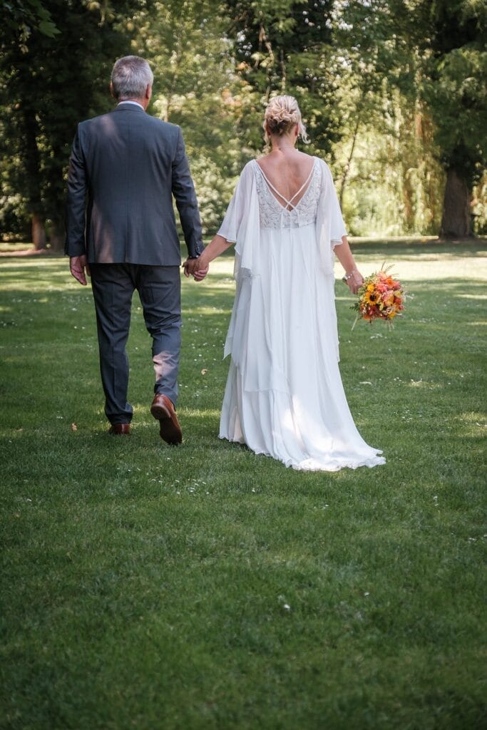 Hochzeitskrone Ein Paar geht Hand in Hand über eine Wiese; die Frau trägt ein weißes Kleid und hält einen Blumenstrauß in der Hand. So werden echte Momente eingefangen. Diese bildschöne Szene ist genau das, was man von natürlichen Hochzeitsfotos eines talentierten Hochzeitsfotografen aus Wien und Niederösterreich erwartet.