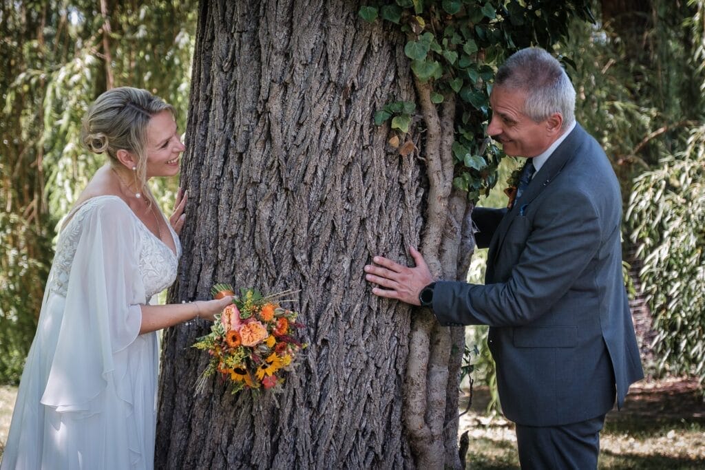 Hochzeitskrone Braut und Bräutigam stehen auf beiden Seiten eines Baumes und tauschen einen Blick, der echte Momente einfängt. Die Braut hält einen Strauß gelber und orangefarbener Blumen vor dem üppigen grünen Laub und schafft so authentische Hochzeitsfotos, die für immer in Erinnerung bleiben.