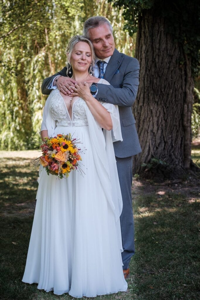 Hochzeitskrone Ein Paar steht im Freien in Hochzeitskleidung, festgehalten in authentischen Hochzeitsfotos. Die Braut hält einen Strauß gelber und orangefarbener Blumen, während der Bräutigam sie von hinten mit geschlossenen Augen umarmt, neben einem Baum. Diese natürlichen Hochzeitsfotos strahlen echte Wärme und Liebe aus.