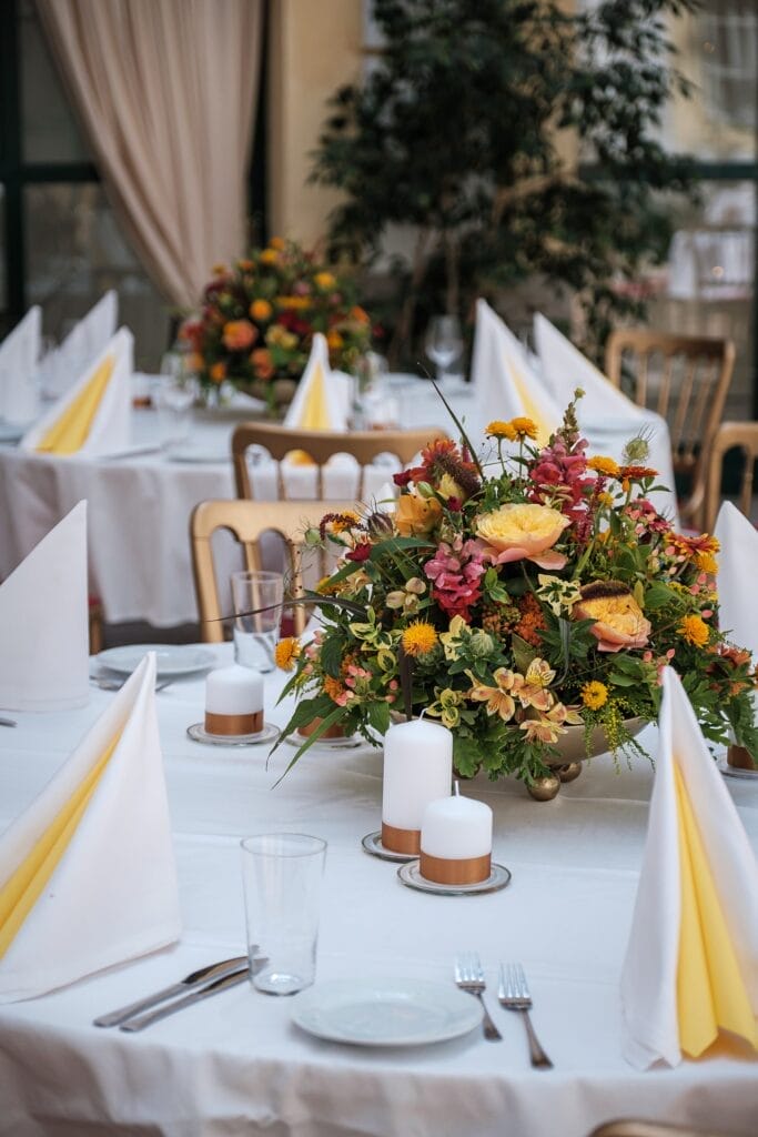 Hochzeitskrone Elegantes Esszimmer-Setup mit weißen Tischdecken, gefalteten gelben und weißen Servietten, floralen Mittelstücken und Kerzen fängt echte Momente ein, perfekt für natürliche Hochzeitsfotos von einem talentierten Hochzeitsfotografen in Wien und Niederösterreich.