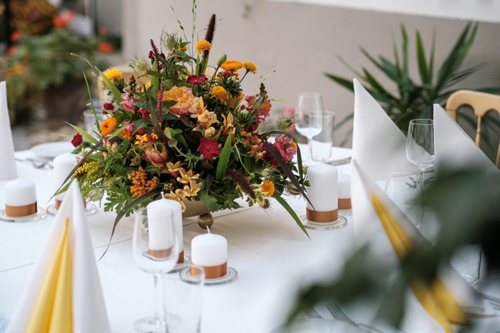 Hochzeitskrone Das farbenfrohe Blumengesteck, flankiert von weißen Kerzen und gelben Servietten auf einem sauberen Tisch, ruft echte Momente hervor, die an authentische Hochzeitsfotos erinnern.