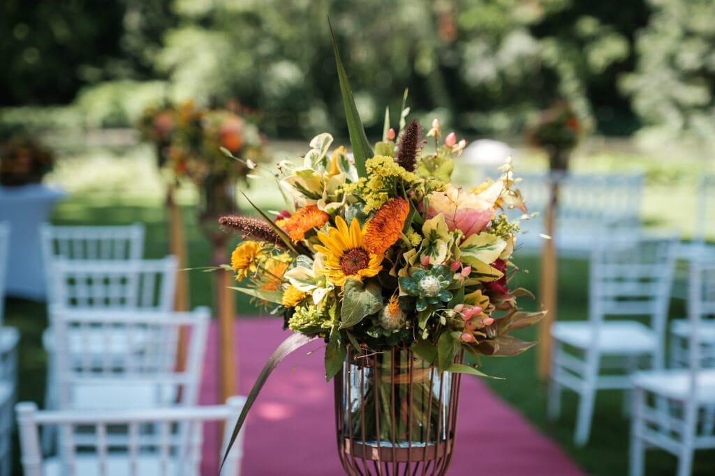 Hochzeitskrone Nahaufnahme eines farbenfrohen Blumenarrangements mit Sonnenblumen und verschiedenen Blüten auf einem Metallständer. Weiße Stühle und ein roter Teppich säumen den Hintergrund in einer Außenkulisse und fangen echte Momente ein – perfekt für einen authentischen Hochzeitsfoto-Touch von einem erfahrenen Hochzeitsfotografen in Wien und Niederösterreich.