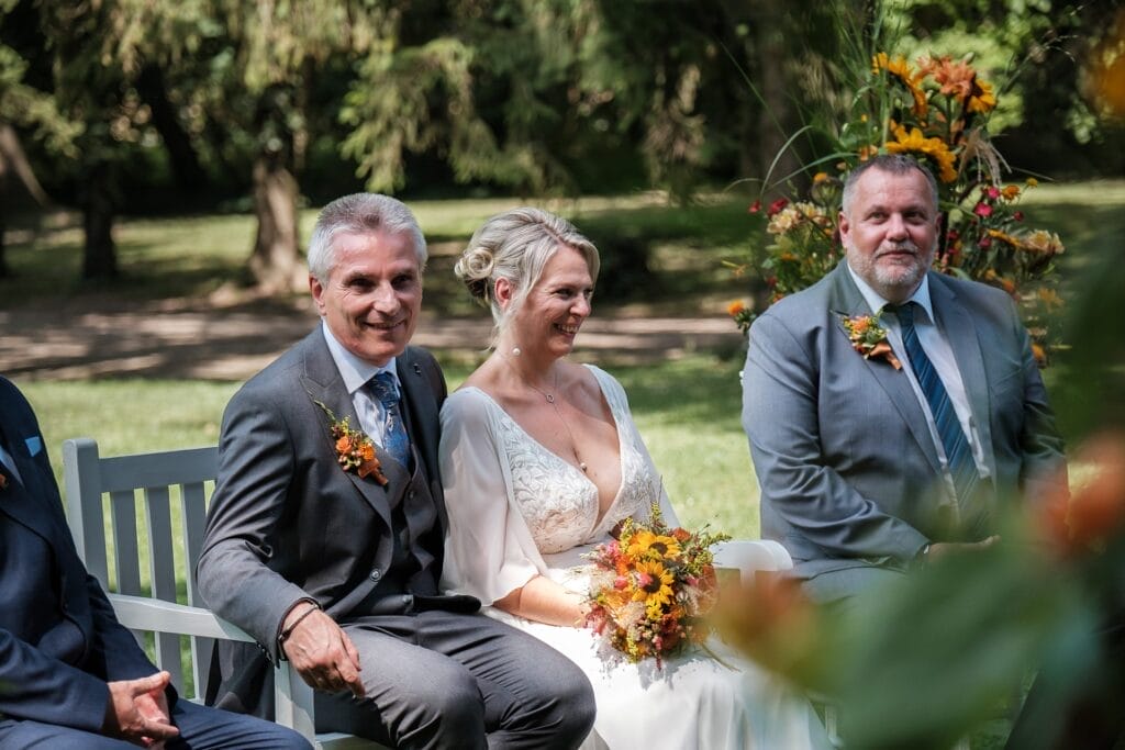 Hochzeitskrone Braut und Bräutigam sitzen mit einem weiteren Mann im Freien auf einer Bank, umgeben von Grün und Blumenarrangements, und fangen die Essenz authentischer Hochzeitsfotos ein.