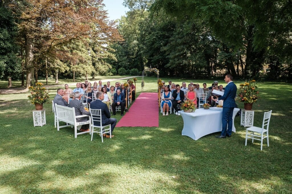 Hochzeitskrone Eine Hochzeitszeremonie im Freien findet statt, die Gäste sitzen auf weißen Stühlen, ein roter Teppich führt zum Trauzeugen unter den Bäumen. Diese echten Momente werden von einem Hochzeitsfotografen in Wien und Niederösterreich festgehalten und sorgen für authentische Hochzeitsfotos, die jedes geschätzte Detail bewahren.