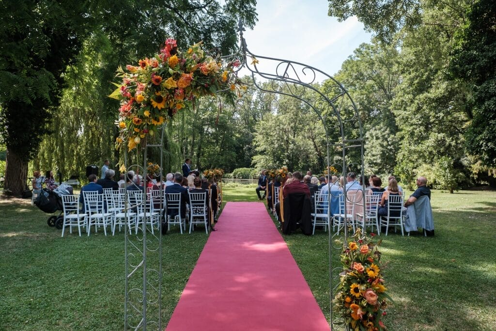 Hochzeitskrone Hochzeitszeremonie im Freien mit Gästen auf weißen Stühlen. Ein roter Teppich führt zu einem Blumenbogen, der authentische Hochzeitsfotos einfängt. Bäume umgeben die Szene und verstärken den Charme echter Momente.