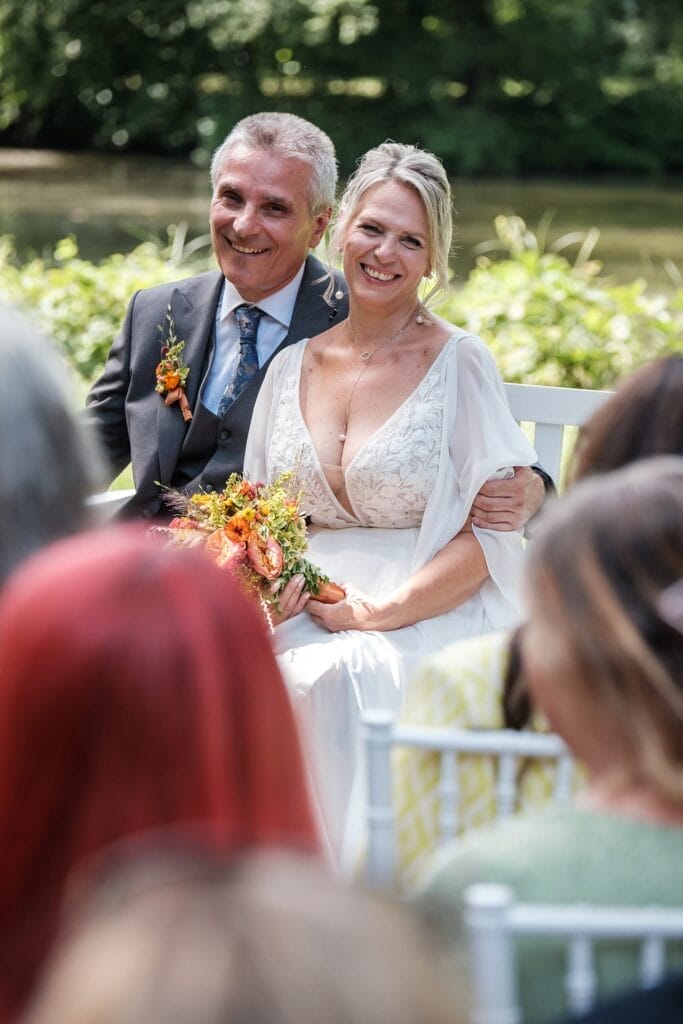 Hochzeitskrone Eine lächelnde Braut und ein Bräutigam sitzen während einer Hochzeitszeremonie im Freien zusammen. Die Braut hält einen Strauß orangefarbener und gelber Blumen in der Hand und fängt damit die Essenz authentischer Hochzeitsfotos ein, die ihre Freude auf natürliche Weise widerspiegeln.