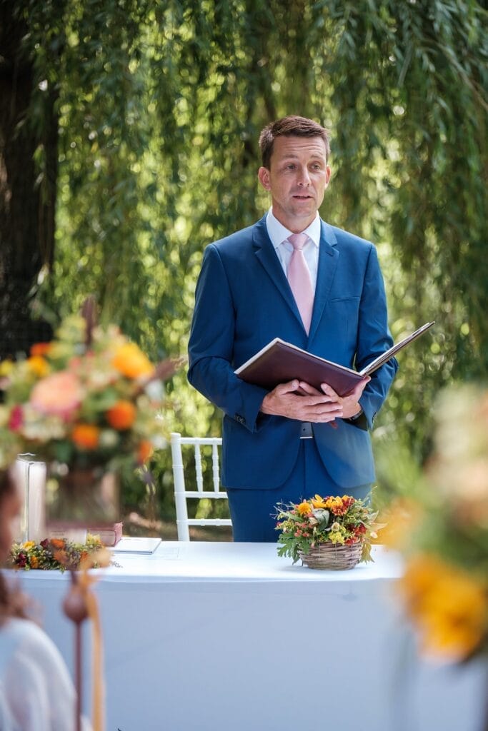 Hochzeitskrone Ein Mann in blauem Anzug und rosa Krawatte steht im Freien und hält ein offenes Buch in der Hand. So entstehen echte Momente. Im Vordergrund verstärkt ein mit Blumenarrangements geschmückter Tisch den Charme der Szene.