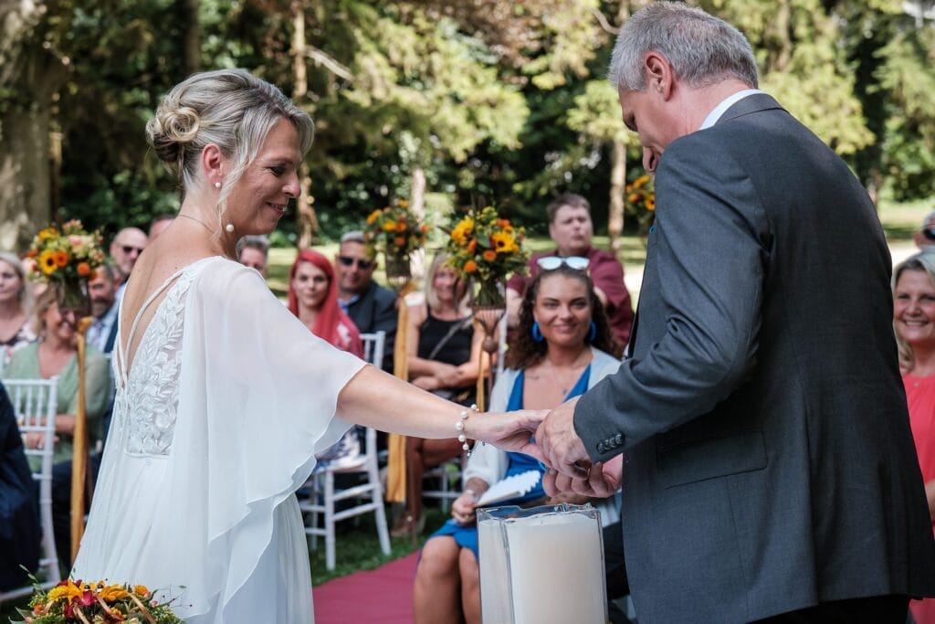 Hochzeitskrone Ein Paar steht während einer Hochzeitszeremonie im Freien Händchen haltend zusammen, umgeben von sitzenden Gästen und mit Blumen geschmückt, festgehalten von einem Hochzeitsfotografen in Wien und Niederösterreich für natürliche und authentische Hochzeitsfotos.