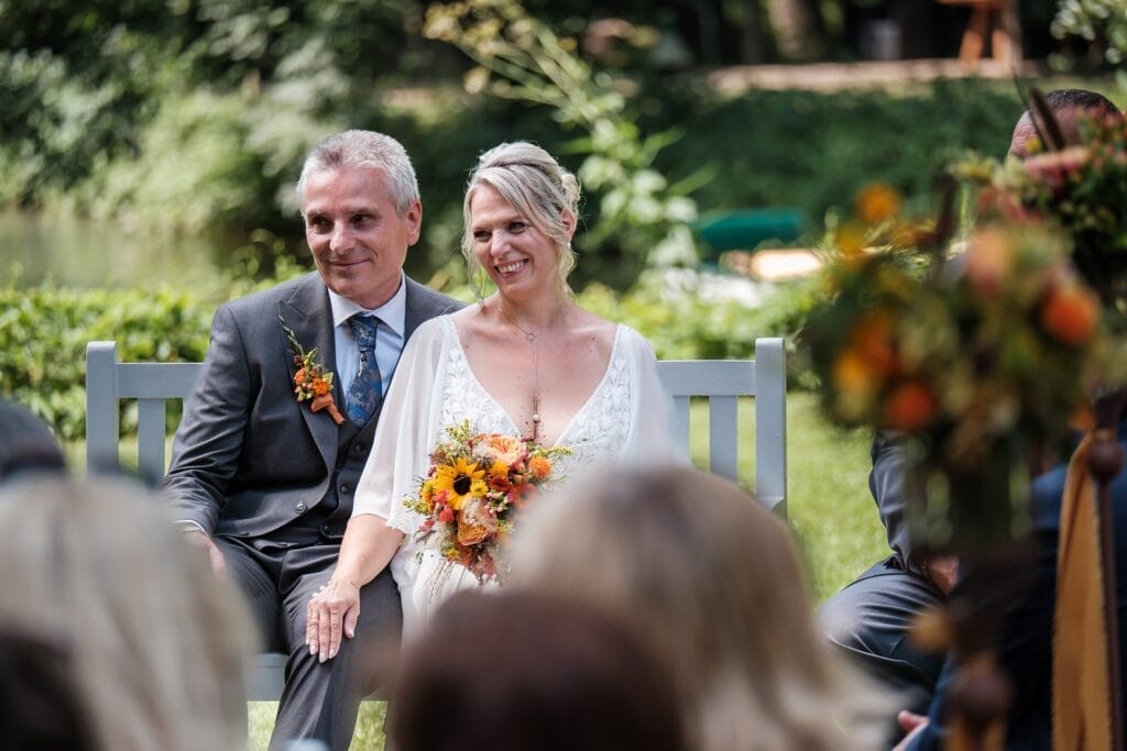 Hochzeitskrone Ein Paar sitzt während einer Hochzeitszeremonie zusammen auf einer Bank im Freien. Die Frau, die einen Strauß Sonnenblumen umklammert, verleiht diesem Moment Charme, festgehalten von einem Hochzeitsfotografen in Wien und Niederösterreich, der auf natürliche Hochzeitsfotos spezialisiert ist.