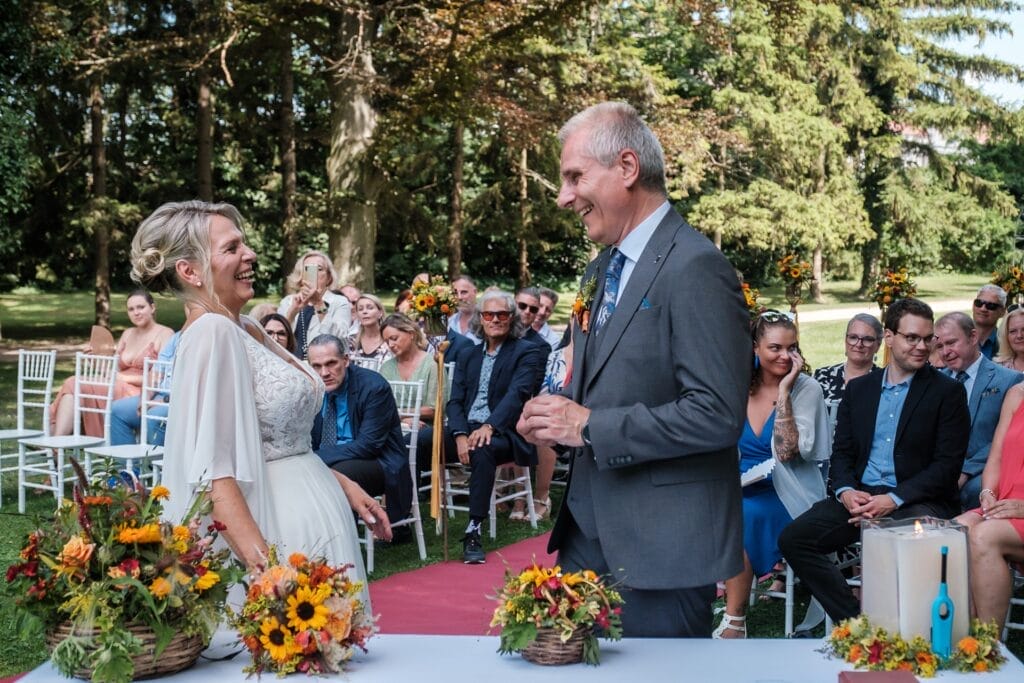 Hochzeitskrone Ein Paar lächelt sich während einer Hochzeitszeremonie im Freien an, umgeben von Gästen, die auf weißen Stühlen sitzen – und fängt echte Momente durch natürliche Hochzeitsfotos ein, aufgenommen von einem Hochzeitsfotografen in Wien und Niederösterreich.