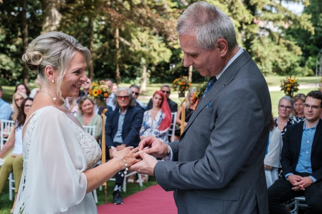 Hochzeitskrone Ein Paar tauscht während einer Hochzeitszeremonie im Freien Ringe aus, wunderschön festgehalten von einem Hochzeitsfotografen in Wien und Niederösterreich, mit im Hintergrund sitzenden Gästen.