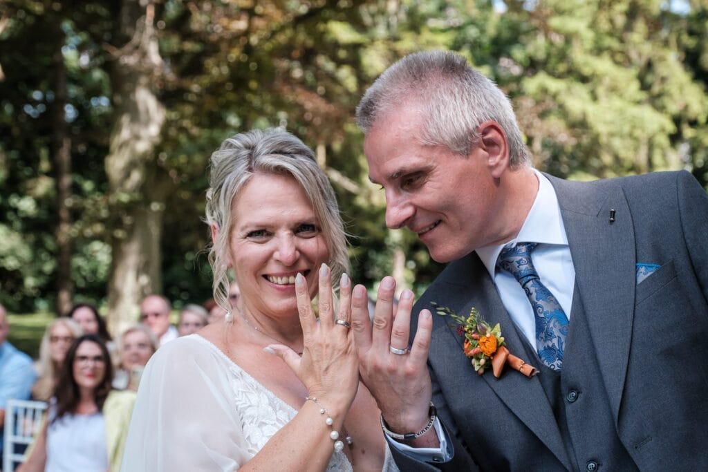 Hochzeitskrone Ein Paar lächelt und zeigt seine Ringe inmitten einer malerischen Naturkulisse in Niederösterreich, mit Gästen im Hintergrund, die bezaubernde Momente purer Freude und Liebe einfangen.