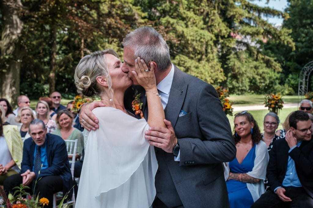 Hochzeitskrone Bei einer Hochzeitszeremonie im Freien küsst sich ein Paar „echte Momente“, umgeben von in der Nähe sitzenden Gästen und mit Bäumen im Hintergrund.